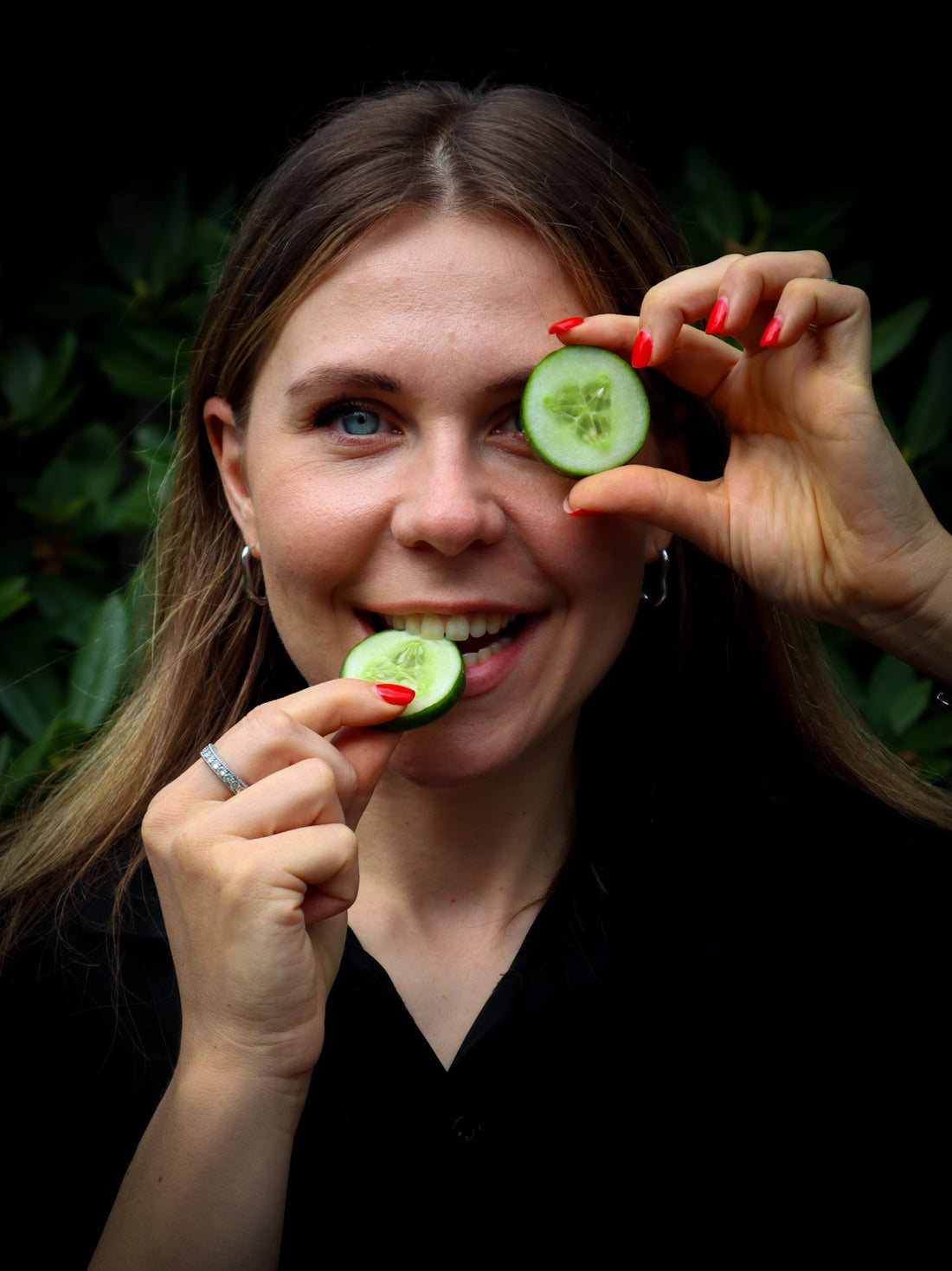 A woman is smiling, holding one cucumber slice near her eye and biting into another.